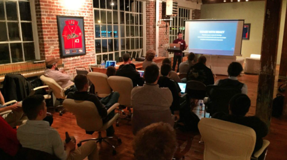 A group of people sit and listen to a speaker at the front of the room.